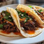 Three soft corn tortillas filled with shredded Crockpot Korean Beef Tacos, topped with cilantro, onions, and spicy orange sauce.