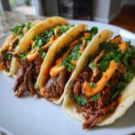 Four fully loaded Crockpot Korean Beef Tacos filled with shredded beef, cilantro, and a spicy orange sauce on a white plate.