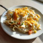 A spoonful of creamy Dump and Bake Chicken Casserole lifted from a plate, showing shredded chicken, pasta, vegetables, and melted cheddar cheese.
