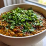 A close-up of a white bowl filled with rich, orange-brown Easy Green Lentil Dal, topped generously with fresh chopped cilantro.