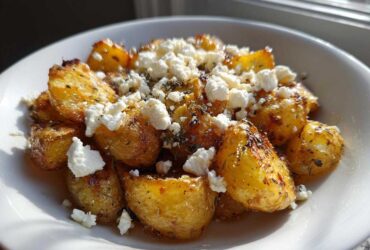 Close-up of golden, roasted potatoes topped with crumbled feta cheese and herbs, representing Greek Feta Roast Potatoes.