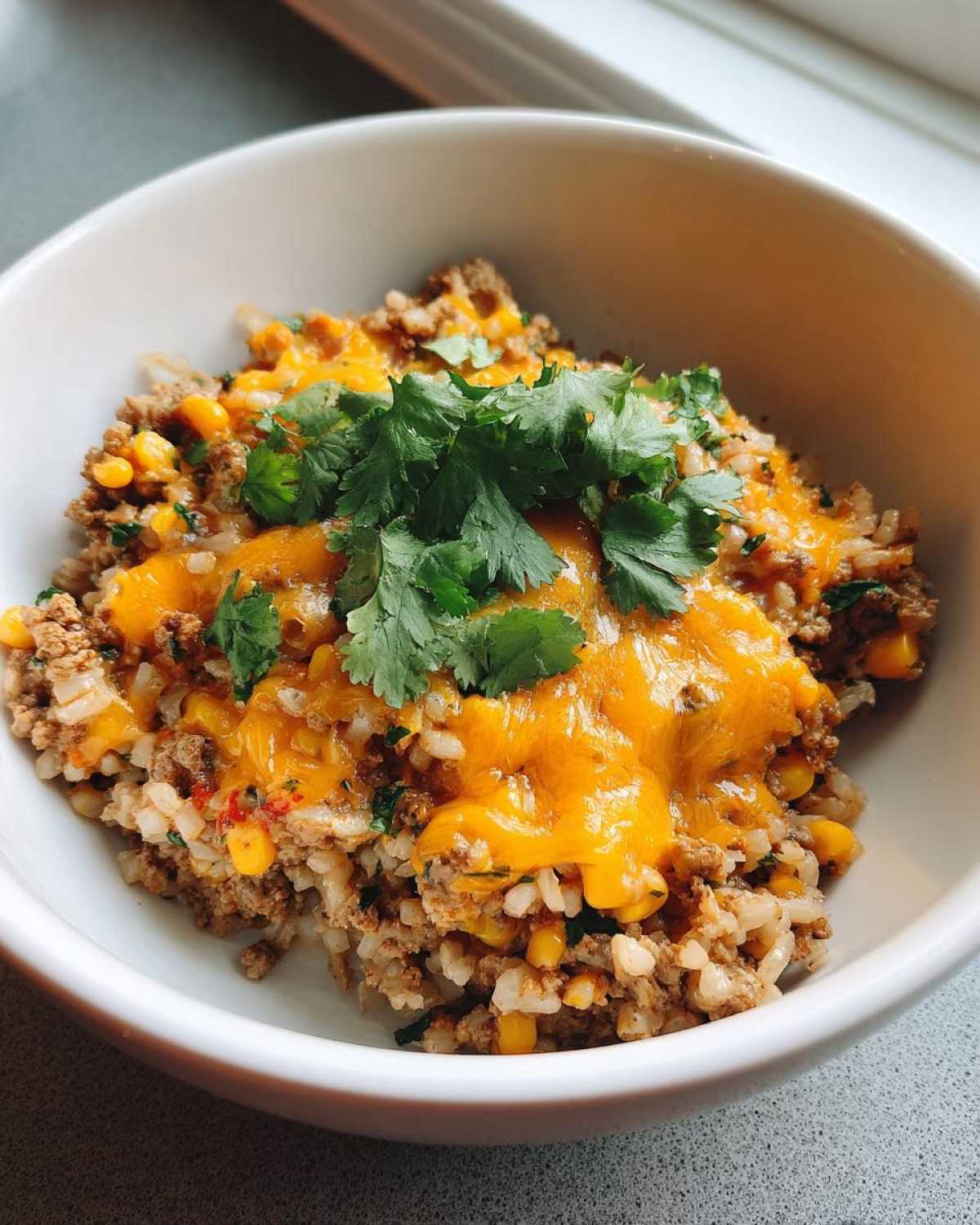 Close-up of a white bowl filled with Ground Turkey Rice Bowls, topped with melted cheddar cheese and fresh cilantro.