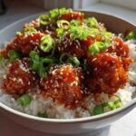 A close-up of a bowl filled with white rice topped with sticky, glazed Honey Sriracha Chicken, garnished with sesame seeds and sliced green onions.