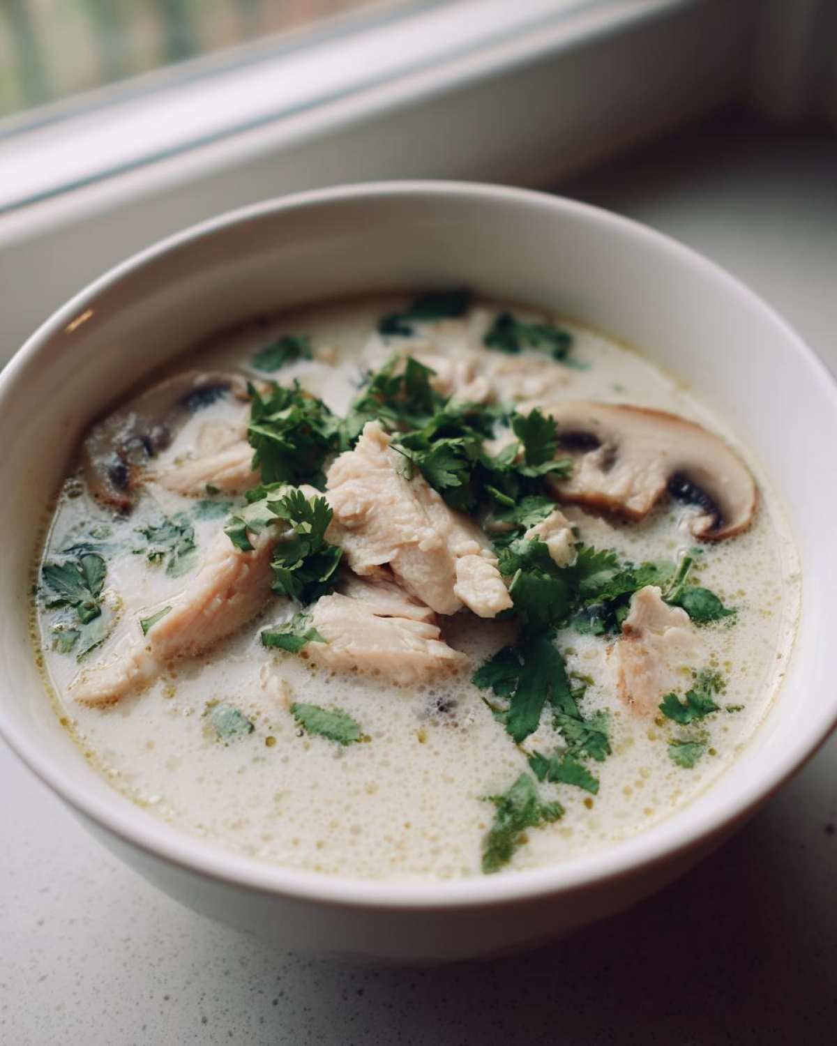 Close-up of Irresistible Thai Chicken Soup in a white bowl, topped with shredded chicken, mushrooms, and fresh cilantro.
