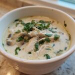 Close-up of a white bowl filled with Irresistible Thai Chicken Soup, featuring shredded chicken, mushrooms, and fresh cilantro garnish.