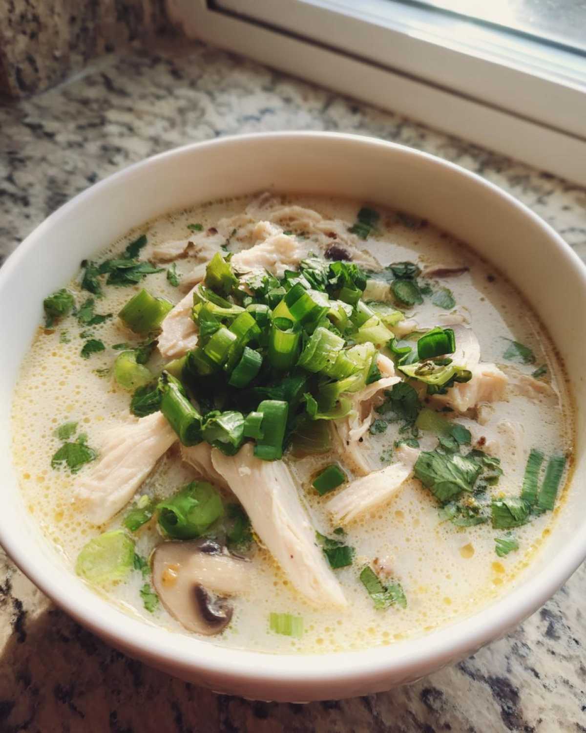 A close-up of a white bowl filled with creamy Irresistible Thai Chicken Soup, topped with shredded chicken, mushrooms, and fresh green onions.