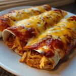 Three rolled Joanna Gaines Enchiladas covered in red sauce and melted cheddar and Monterey Jack cheese on a white plate.
