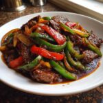A close-up of a white bowl filled with glossy Keto Beef Pepper Steak mixed with bright red and green bell peppers.
