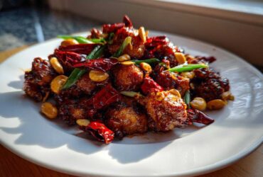 A close-up of Keto Kung Pao Chicken served on a white plate, featuring glossy chicken pieces, dried red chilies, and peanuts.