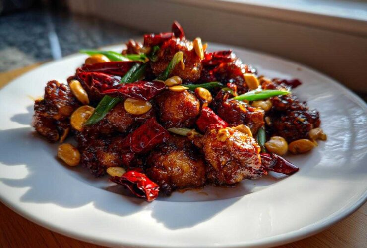 A close-up of Keto Kung Pao Chicken served on a white plate, featuring glossy chicken pieces, dried red chilies, and peanuts.