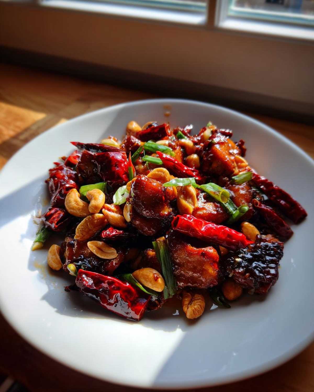 A close-up of Keto Kung Pao Chicken served on a white plate, featuring glazed chicken, whole dried chilies, cashews, and green onions.