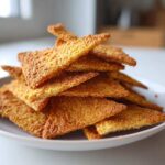 A close-up stack of golden brown, triangular Keto Sour Cream Crackers piled high on a white plate.