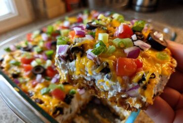A hand lifts a square serving of the layered dip, showing creamy layers, melted cheese, and colorful toppings on this Avocado Party Dip.