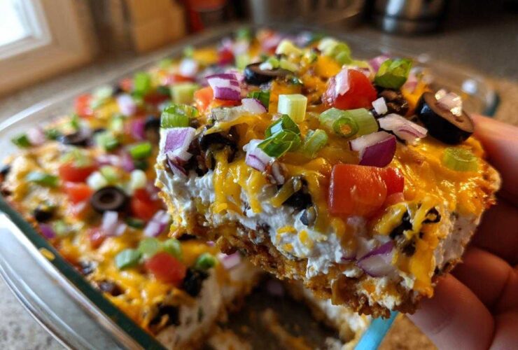 A hand lifts a square serving of the layered dip, showing creamy layers, melted cheese, and colorful toppings on this Avocado Party Dip.