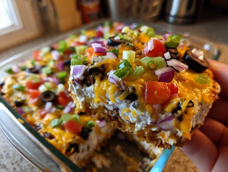 A hand lifts a square serving of the layered dip, showing creamy layers, melted cheese, and colorful toppings on this Avocado Party Dip.