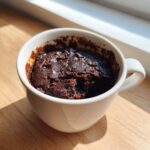 A rich, dark chocolate Single Serve Protein Brownie baked inside a white mug, sitting on a wooden surface.