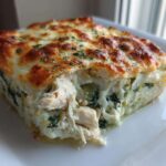 A thick, square slice of Spinach Artichoke Chicken Casserole showing shredded chicken, creamy spinach, and a bubbly, browned cheese crust.