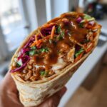 A hand holds a tightly wrapped tortilla filled with shredded chicken, colorful slaw, and drizzled heavily with peanut sauce for a Thai Chicken Wrap Recipe.