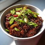 A close-up of a bowl filled with rich, dark Vegan Korean Bulgogi Lentils, topped with green onions and sesame seeds.