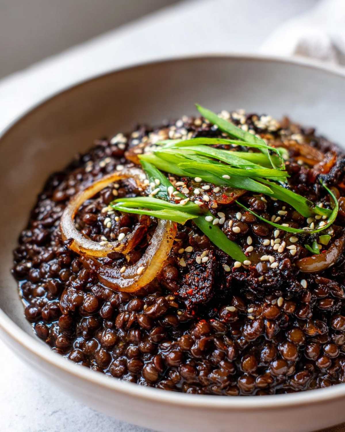 Close-up of rich, dark Vegan Korean Bulgogi Lentils topped with caramelized onions, green onions, and sesame seeds.