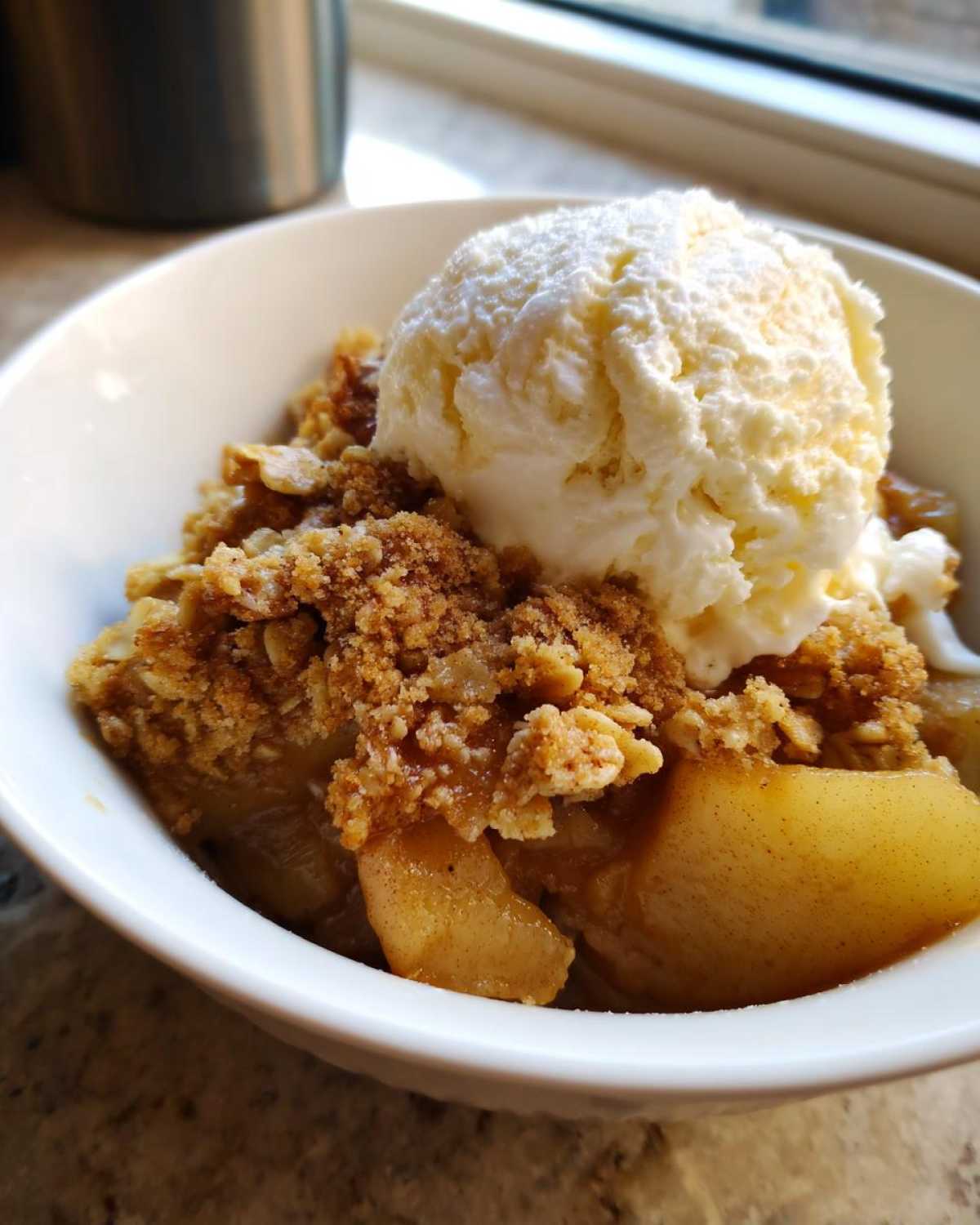 Close-up of a warm serving of Apple Crisp topped with a scoop of vanilla ice cream in a white bowl.