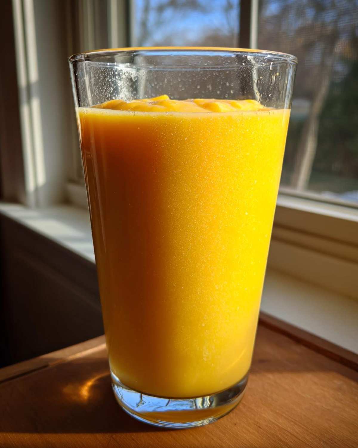 A tall glass filled with a vibrant, thick Banana Mango Smoothie, backlit by a window.