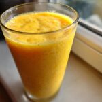 Close-up of a vibrant, thick Banana Mango Smoothie filling a tall glass, sitting near a window.