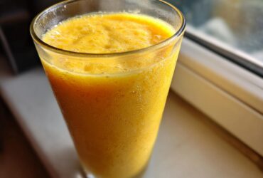 Close-up of a vibrant, thick Banana Mango Smoothie filling a tall glass, sitting near a window.