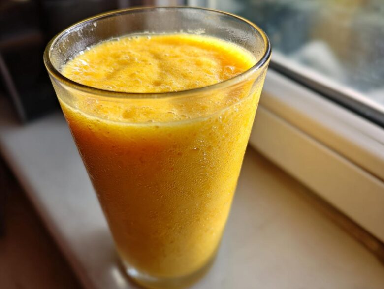 Close-up of a vibrant, thick Banana Mango Smoothie filling a tall glass, sitting near a window.
