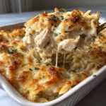 A spoonful of creamy Chicken Orzo Bake being lifted from a white baking dish, showing melted cheese and shredded chicken.
