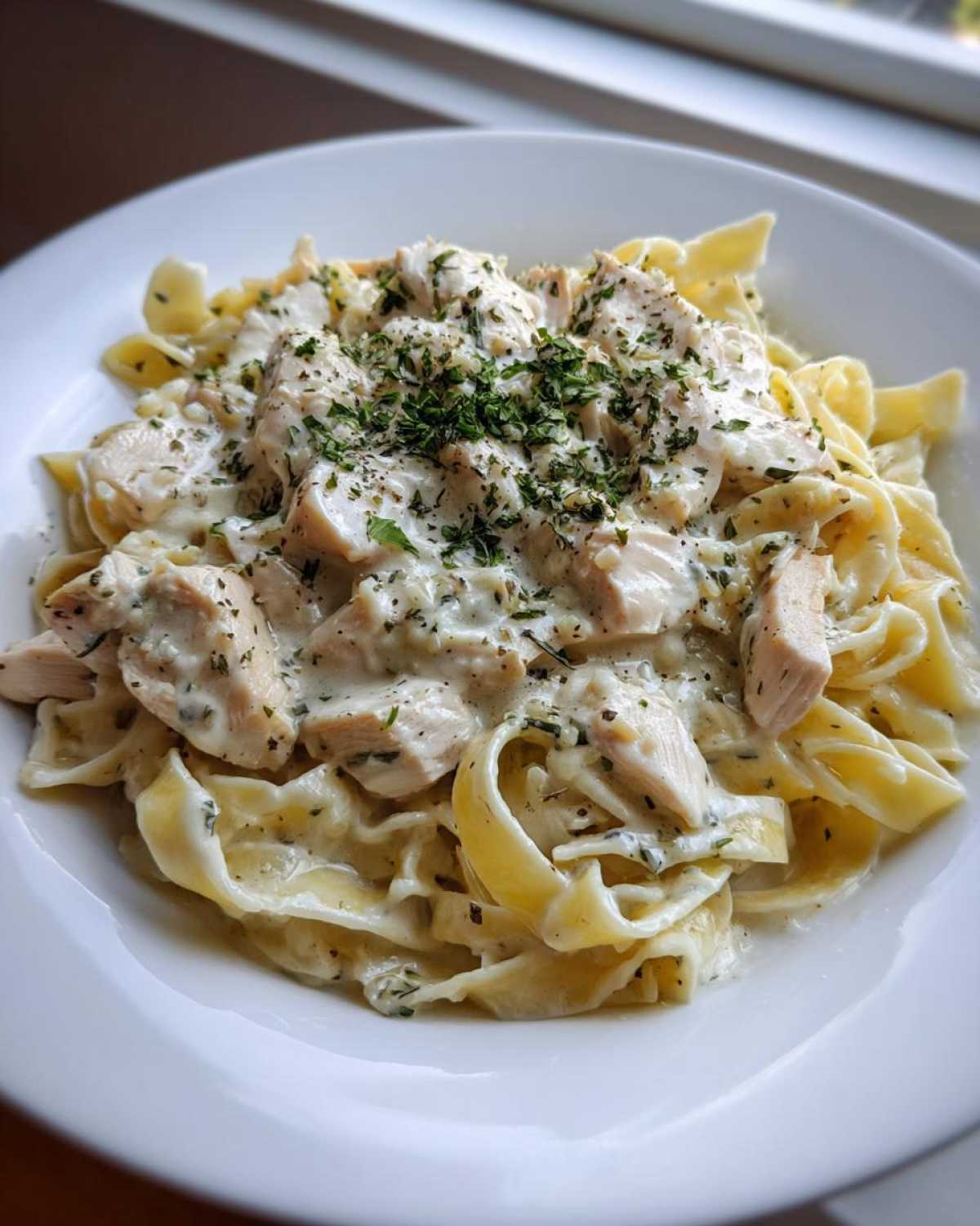 A close-up of a white bowl filled with Creamy Chicken Fettuccine Alfredo, topped with chopped parsley.