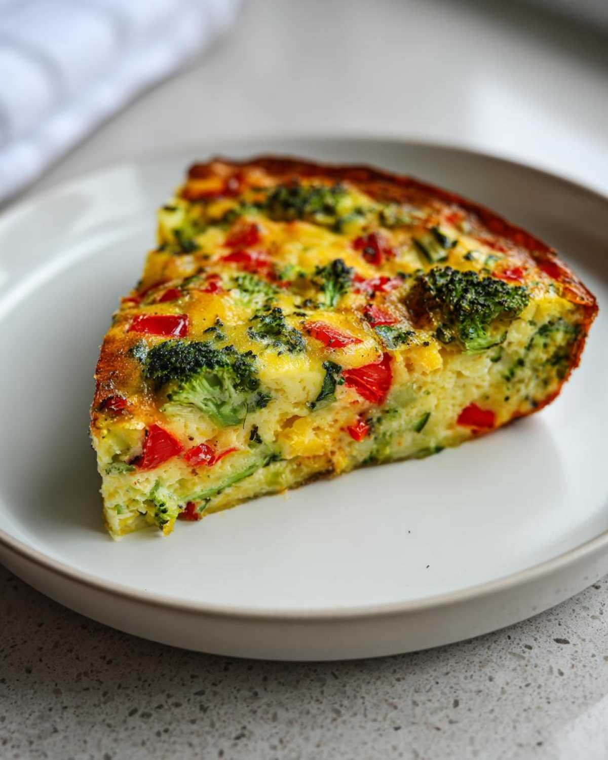 A single slice of Crustless Garden Vegetable Quiche featuring visible broccoli florets and red peppers on a white plate.
