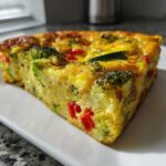 A thick, golden slice of Crustless Garden Vegetable Quiche filled with broccoli, zucchini, and red peppers, served on a white rectangular plate.