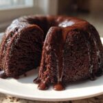 A dark, moist Death By Chocolate Bundt Cake with thick chocolate glaze dripping down the sides, served on a white plate.