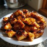 A close-up of glistening Honey Garlic Chicken Bites piled high on a white plate, coated in a dark, sticky glaze and sprinkled with sesame seeds.