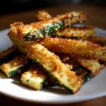 A close-up stack of golden brown, crispy Baked Parmesan Zucchini sticks served on a white plate.