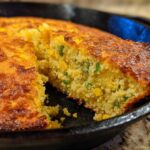 Close-up of a slice being removed from a golden-brown Cast Iron Skillet Cornbread studded with corn and green jalapeño pieces.