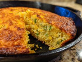 Close-up of a slice being removed from a golden-brown Cast Iron Skillet Cornbread studded with corn and green jalapeƱo pieces.