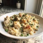 A close-up of a white bowl filled with creamy chicken and rice mixed with bright green peas, topped with melted cheese and parsley.