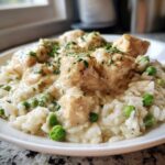 A close-up of a white plate filled with creamy chicken and rice mixed with bright green peas, garnished with parsley.
