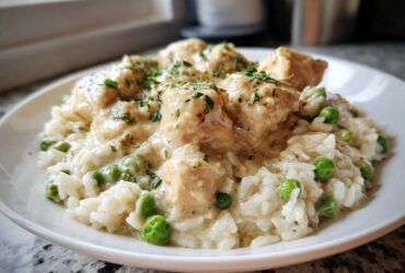 A close-up of a white plate filled with creamy chicken and rice mixed with bright green peas, garnished with parsley.