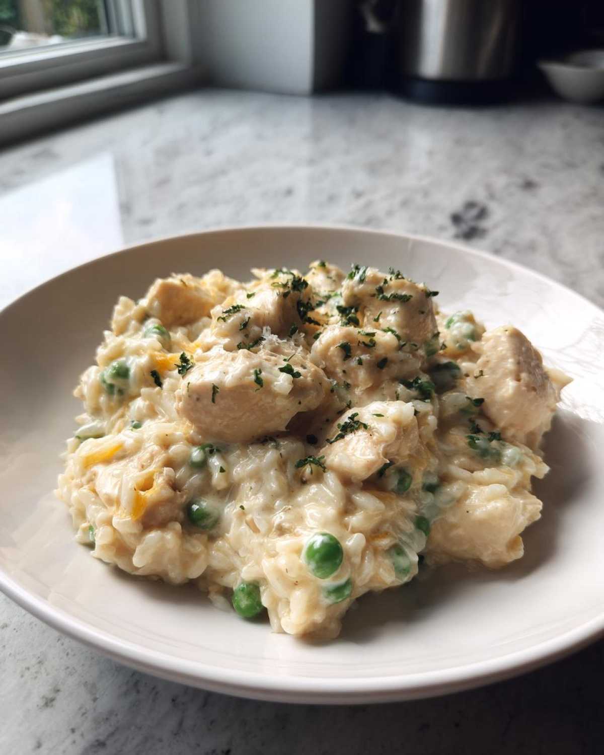 A close-up of a bowl filled with creamy chicken and rice mixed with bright green peas, garnished with parsley.