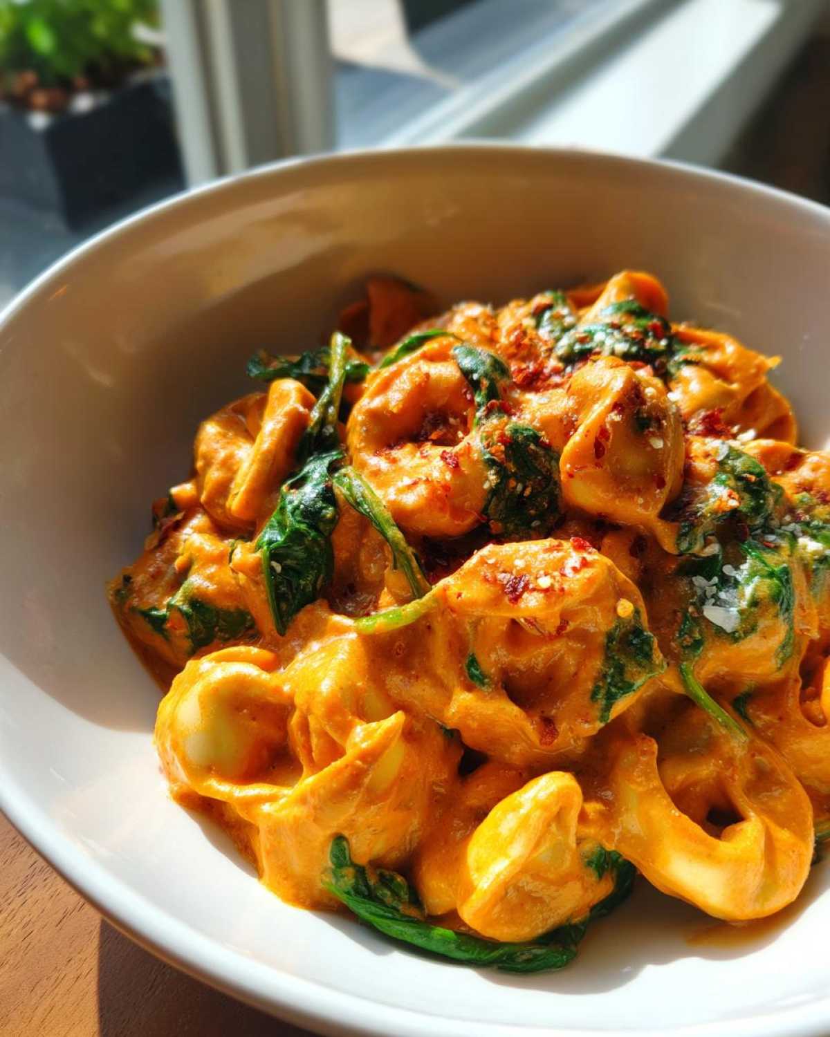 Close-up of a bowl of Marry Me Tortellini coated in a rich, creamy orange sauce with wilted spinach and red pepper flakes.