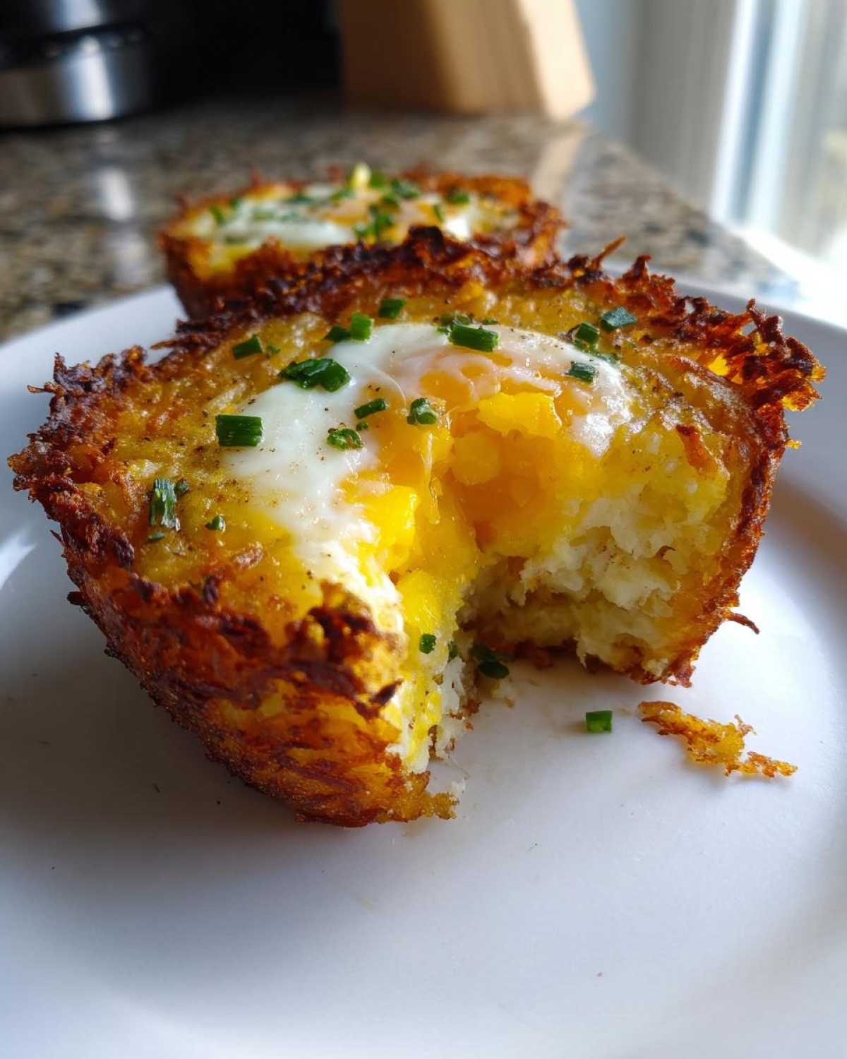 A close-up of a crispy Hash Brown Breakfast Cup with a runny egg yolk spilling out, garnished with chives.
