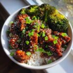 Close-up of a Honey BBQ Chicken Rice Bowl topped with sesame seeds and green onions, served with roasted broccoli florets.