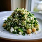 A mound of Jennifer Aniston Salad featuring quinoa, chopped cucumber, feta cheese, chickpeas, and parsley on a white plate.