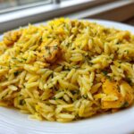 A close-up of a white plate filled with bright yellow Lemon Chicken Orzo Stir Fry, garnished with fresh green herbs.