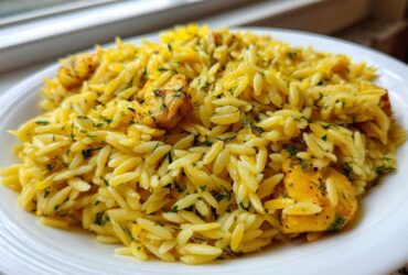 A close-up of a white plate filled with bright yellow Lemon Chicken Orzo Stir Fry, garnished with fresh green herbs.