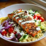Close-up of a seasoned salmon fillet served over quinoa with cucumbers, tomatoes, feta, and tzatziki in a Mediterranean Salmon Bowl.
