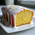 A close-up of a moist Greek Yogurt Lemon Loaf Cake drizzled heavily with white lemon glaze and topped with zest.